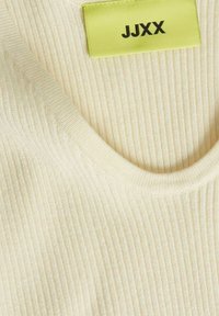 Cremefarbenes, geripptes Strickmaterial mit rundem Ausschnitt und einem limonengrünen Etikett, auf dem der Markenname "JJXX" steht.
