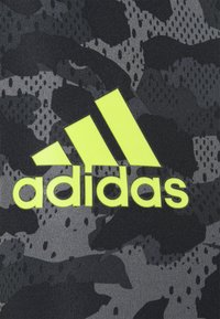 Černá a šedá tkanina s maskáčovým vzorem, která obsahuje velké, jasně žluté logo Adidas v tučném, moderním písmu.