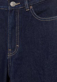 Gros plan sur un jean en denim bleu foncé montrant la poche avant, la boucle de ceinture, le bouton métallique et les détails de couture contrastante.