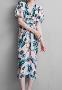 Mujer con un vestido midi floral en tonos pastel y mangas abullonadas, de pie contra una pared gris texturizada ajustándose la manga con la mano.