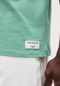 Πράσινο t-shirt Björn Borg με ετικέτα λογότυπου πάνω από λευκό παντελόνι, φορεμένο από ένα άτομο με σκούρο δέρμα ορατό στο χέρι και τον καρπό.