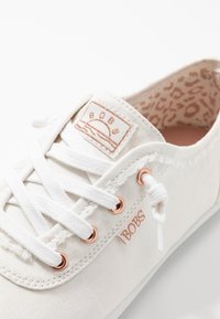 Chaussure en toile blanche avec œillets en cuivre, lacets blancs et patch logo cousu. L'intérieur présente un motif discret imprimé léopard.