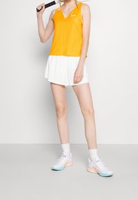Top naranja sin mangas con escote en V, combinado con pantalones cortos blancos. La modelo lleva zapatillas blancas con acentos en tonos pastel. Raqueta de tenis sostenida en la mano derecha.