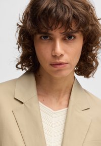 Beige blazer dengan kerah tajam, dilapisi di atas atasan putih bertekstur. Rambut coklat keriting pendek membingkai wajah. Latar belakang netral.