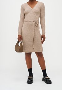 Robe en maille côtelée beige avec un design portefeuille, manches longues et taille ceinturée. Mocassins à plateforme noirs et petit sac à main marron complètent le look.