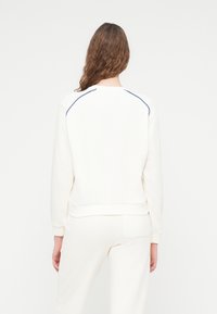 Sweat-shirt blanc avec manches raglan et couture contrastée bleu marine aux épaules. Comprend des poignets et un ourlet côtelés.