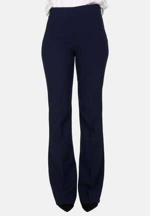 Persona che indossa pantaloni eleganti blu navy scuro a vita alta e una camicia bianca, in piedi di fronte su uno sfondo chiaro.