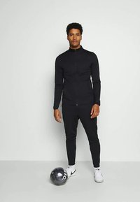 Fato atlético preto com um casaco com fecho éclair e calças justas. Usado com ténis brancos, apresentando uma textura suave e um design minimalista.