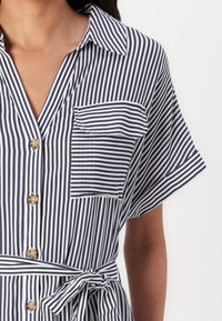 Ριγέ φόρεμα σε ναυτικό και λευκό χρώμα με κοντά μανίκια, κουμπάκι μπροστά, τσέπη στο στήθος και λεπτομέρεια με δέσιμο στη μέση. Διαθέτει μαλακή υφή υφάσματος.