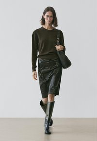 Pull brun foncé, jupe noire brillante, longues bottes noires et sac à main noir texturé. La tenue présente des silhouettes élégantes et modernes.