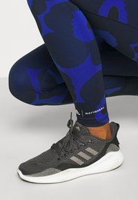 Osoba oblečená do černobílých běžeckých bot Adidas a modrých legín s Marimekko vzorem, která se krčí na šedé podlaze.