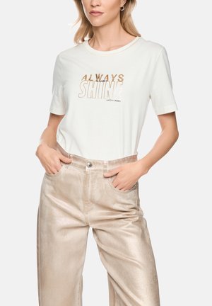 Woman wearing a white short-sleeve shirt with "Always Shine" text and shiny beige high-waisted pants, hands in pockets.