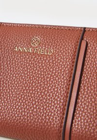 Barna textúrájú tárca arany "ANNA FIELD" logóval. Simának tűnő bőrkiemeléssel és karcsú kialakítással rendelkezik, amely a finom kivitelezést mutatja be.