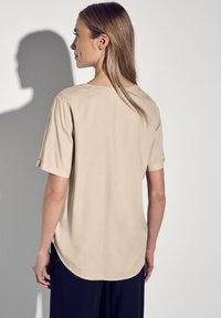 Beige, locker sitzendes Kurzarmshirt mit abgerundetem Saum, Rückenpasse und umgeschlagenen Ärmelbündchen, aus leichtem, glattem Stoff.