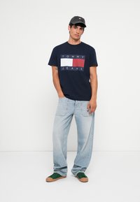 Navyblå bomulds t-shirt med hvid og rød grafisk "TOMMY JEANS" design, sat sammen med lyseblå løse jeans og grønne sneakers.