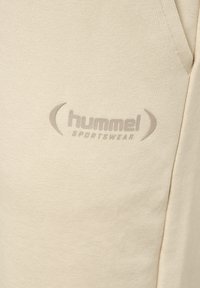 Beige stof met geperst "hummel SPORTSWEAR"-logo en een deel van een naad, waarschijnlijk op sportkleding.