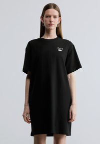 Čierne bavlnené tričko šaty s voľn�ým strihom, krátkymi rukávmi a malým bielym logom na hrudi. Hladká textúra a dĺžka po kolená.