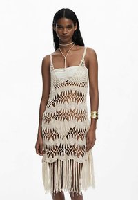 Rochie bej, tricotată, cu model ajurat în formă de valuri și franjuri la tiv. Are bretele subțiri și un design stratificat peste o rochie interioară mulată.