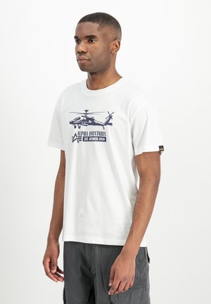 Uomo che indossa una maglietta bianca con grafica di elicottero e scritta "Alpha Industries Est. October 1959", abbinata a pantaloncini cargo scuri.