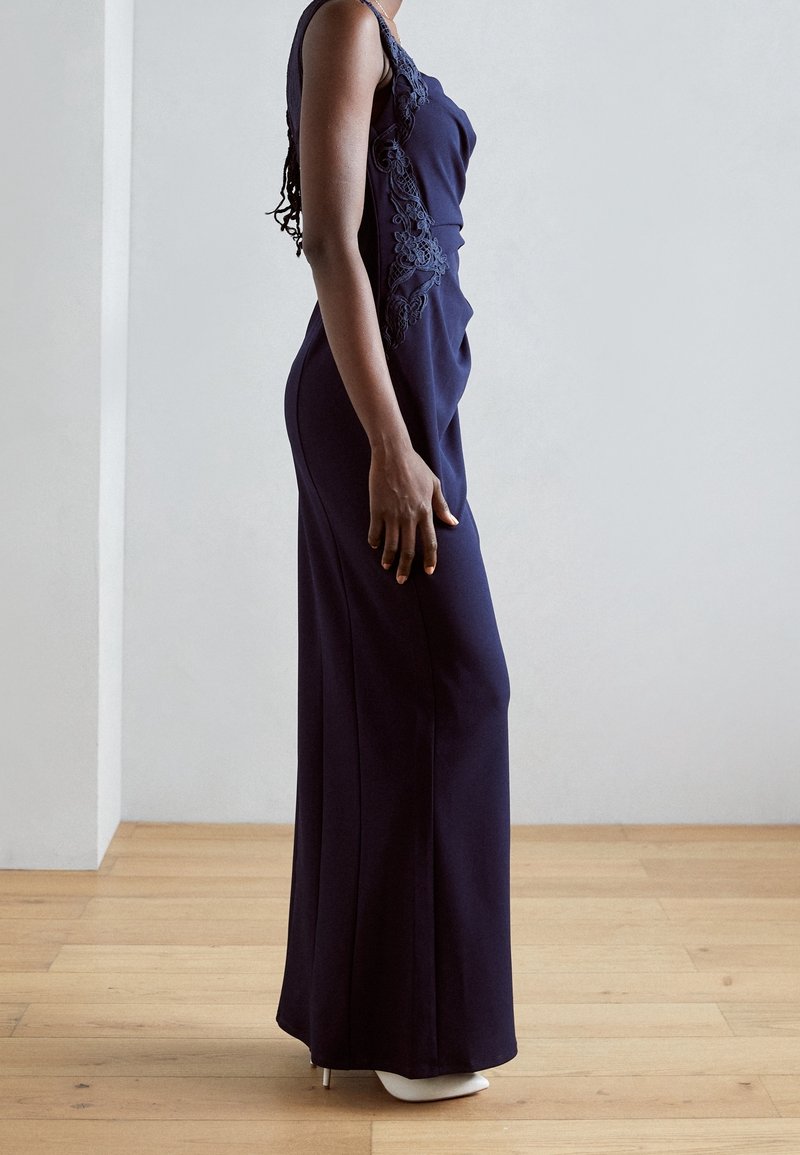 Navyblauwe lange jurk met een aansluitend lijfje en zijdetails van kant. Heeft een slanke silhoutte en een gladde textuur, gecombineerd met witte hoge hakken.