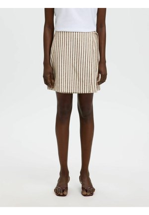 Femme portant une jupe portefeuille beige et marron à rayures verticales, un haut blanc et des sandales ouvertes marron, debout devant un fond uni.