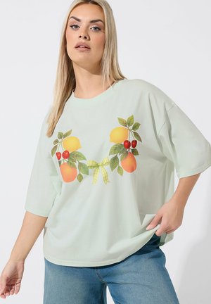 Mujer rubia con camiseta suelta de color verde claro con diseño de frutas y hojas, y vaqueros azules, de pie frente a un fondo liso.