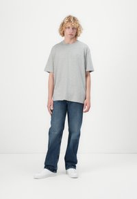 Graues Baumwoll-T-Shirt mit kurzen Ärmeln und rundem Halsausschnitt; ausgestattet mit einem kleinen roten Logo auf der Brust, kombiniert mit blauen Jeans und weißen Sneakers.