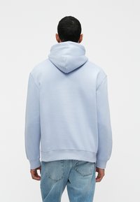 Svetlomodrá mikina s kapucňou s voľným strihom, rebrovanými manžetami a lemom a mäkkou textúrou. Nosená s svetlomodrými džínsami.