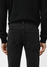 Jeans de mezclilla negra con una silueta ajustada, que cuentan con dos bolsillos traseros y detalles de costura minimalistas. Combinados con un suéter negro.