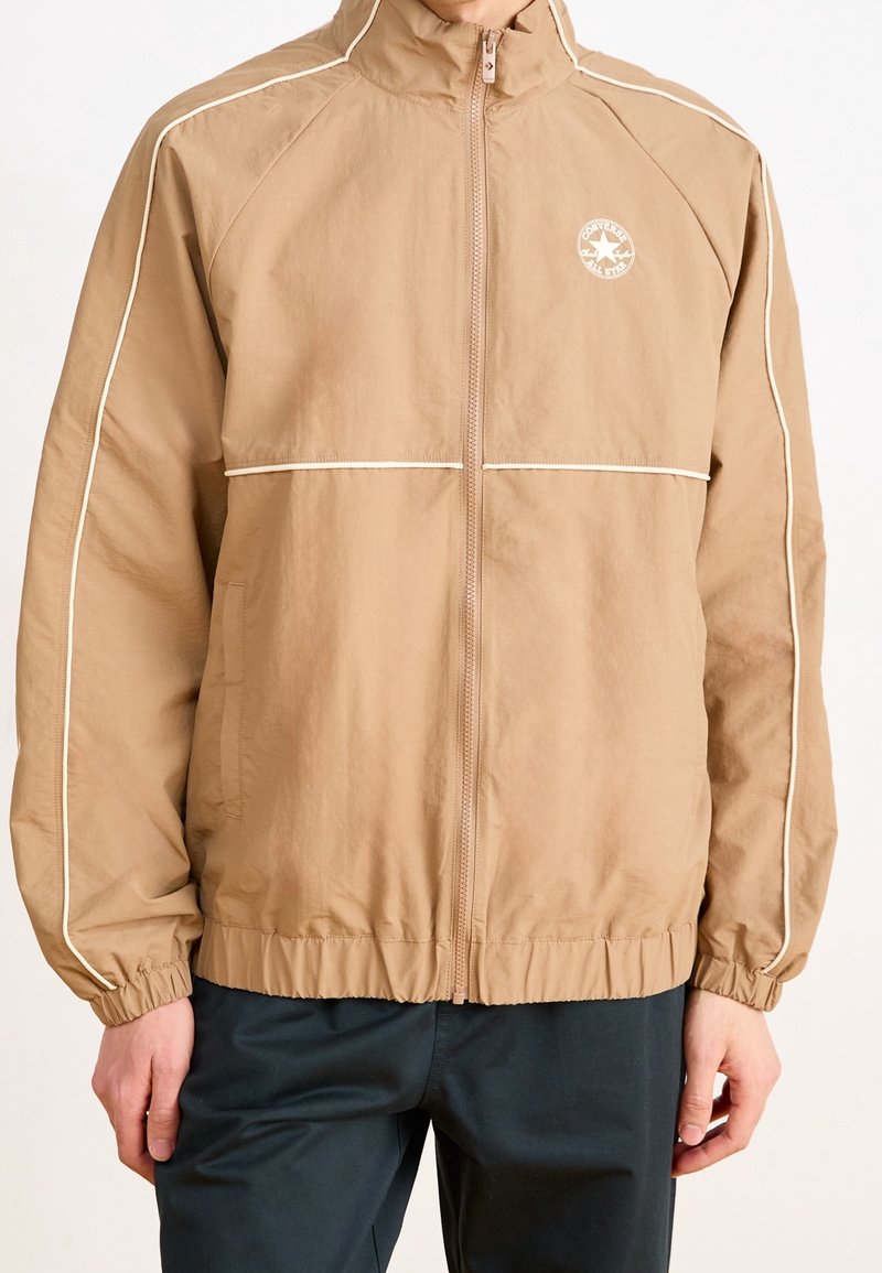 Hellbraune Windjacke mit hochgezogenem Kragen, weißen Paspeln entlang der Nähte, elastischen Bündchen und einem Reißverschluss an der Vorderseite. Mit Logo auf der Brust.