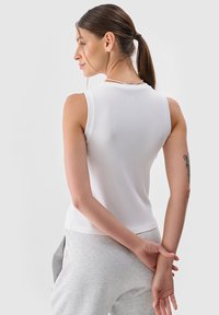 Biała bezrękawnik z prążkowaną teksturą, okrągłym dekoltem i małym logo z tyłu. Stylizowany z szarymi spodniami dresowymi.