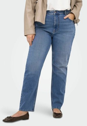 Naine, kes kannab sirge lõikega siniseid teksapükse, beež jakki, valget toppi ja pruune madalaid jalanõusid, seisab ühe käega taskus.