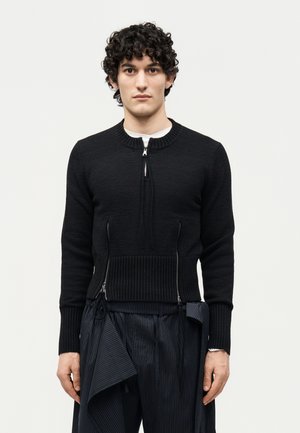 Mann mit lockigem Haar, der einen schwarzen Reißverschluss-Pullover mit geripptem Saum und schwarze Nadelstreifenhose mit Stoffdetails trägt, steht vor einem schlichten Hintergrund.