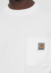 Close-up of person wearing white T-shirt with chest pocket, featuring a gray and gold Carhartt logo patch.