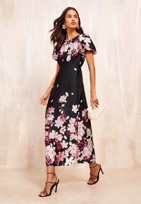 Melnā ziedu maksi kleita ar rozā un baltām ziediem, īsām pūstām piedurknēm un piegulošu augšdaļu. Kombinēta ar melnām siksniņām apaviem un baltu rokassomu.