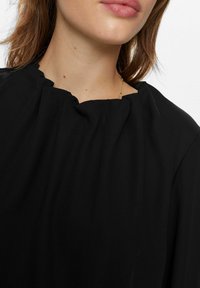 Blusa nera con un collo plissettato texturizzato. Il tessuto appare morbido e liscio, con una vestibilità rilassata e pochi elementi di design.