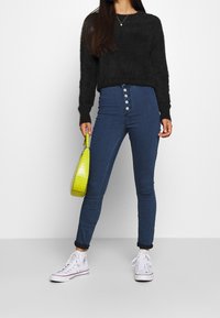 Mujer con suéter de felpa negro, jeans de tiro alto azul con botones frontales, zapatillas deportivas blancas de caña alta, sosteniendo un pequeño bolso de mano amarillo neón.