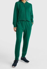 Sweat à capuche zippé vert avec une coupe courte et des poches avant, associé à un pantalon de jogging assorti. Le tissu semble doux et texturé.