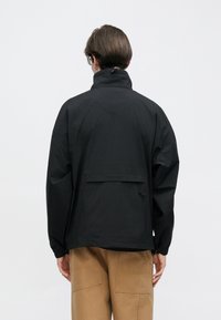 Μαύρο, ελαφρύ μπουφάν με ψηλό γιακά, ελαστικά μανίκια και μια πίσω τσέπη. Το ύφασμα φαίνεται λείο με ματ φινίρισμα.