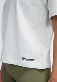 Camiseta gris claro con textura moteada, de manga corta, y un logo negro de "hummel" en el dobladillo izquierdo, combinada con leggings verde oliva.