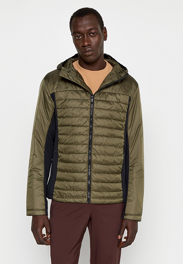 Groene pufferjack met een capuchon, met een gewatteerde textuur en zwarte zijpanelen. Voorzien van een ritssluiting aan de voorkant, gedragen over een lichtbruin shirt.