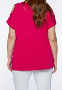 Woman wearing a bright pink cold-shoulder top and white pants, shown from the back with long wavy brown hair.