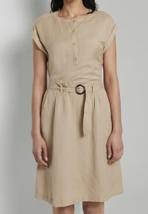 Mujer con un vestido beige de manga corta con escote abotonado y cinturón de tela con hebilla circular en la cintura.
