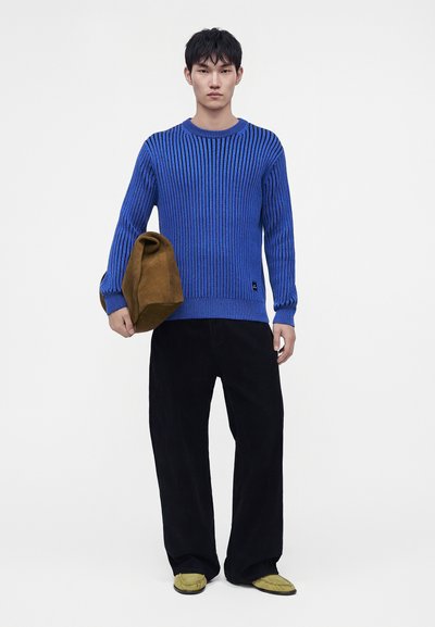 Jeune homme portant un pull côtelé bleu, un pantalon large noir, des chaussures jaunes et tenant un sac marron sur fond blanc.