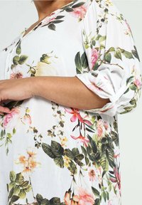 Personne portant un chemisier blanc à motifs floraux roses et verts, avec des manches retroussées et une encolure boutonnée.