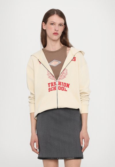 Krémová mikina na zips s červeným potlačou "FASHION SCHOOL", skombinovaná s hnedým topom s výrezom v tvare diamantového tvaru a šedou rebrovanou sukňou.