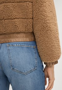 Brązowa puszysta kurtka z teksturowaną powierzchnią i elastycznym paskiem, w połączeniu ze średnioindigo dżinsami z tylną kieszenią.