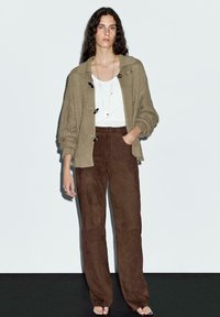 Beige stickad cardigan med stora knappar, bärs över en vit topp och bruna mockabyxor med vida ben, placerad mot en enkel ljus bakgrund.