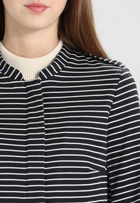 Czarna i biała bluzka w paski z wysokim, kremowym kołnierzem. Miękka w dotyku, dopasowana, z centralnym szwem i subtelnymi detalami przeszyć.