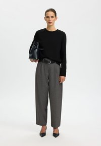 Zwarte top met lange mouwen, grijze op maat gemaakte hoge taillebroek, zwarte riem en zwarte puntige hakken, met een gestructureerde zwarte tas in de hand.